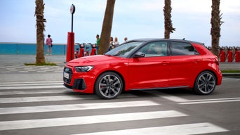 A bright red Audi A1 car crosses a seaside road with blurred palm trees and ocean in the background. Pedestrians stroll on the sidewalk, evoking a relaxed coastal vibe.