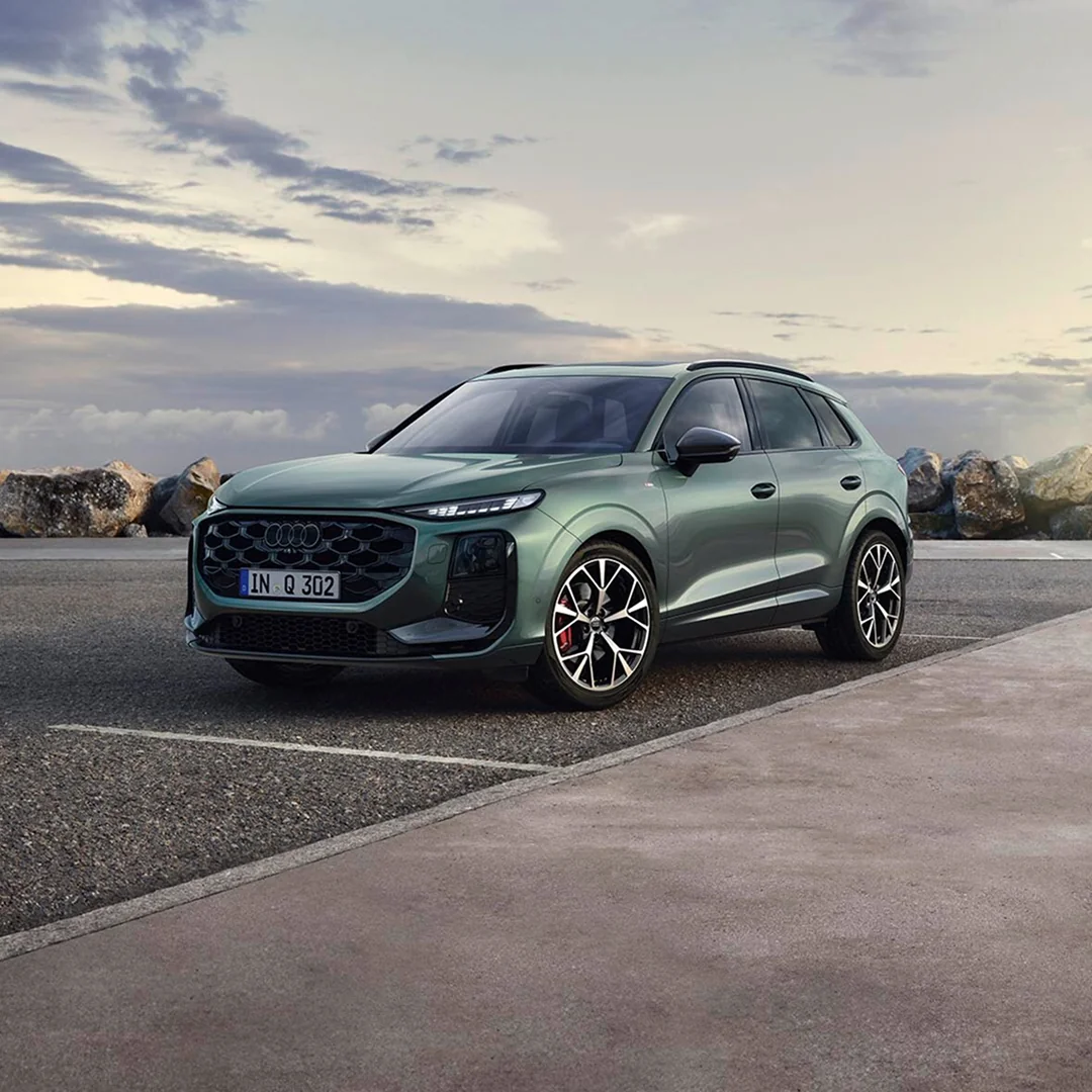 A grey Audi SUV is parked at a charging station beside a modern building. The scene suggests innovation and sustainability against a scenic backdrop.