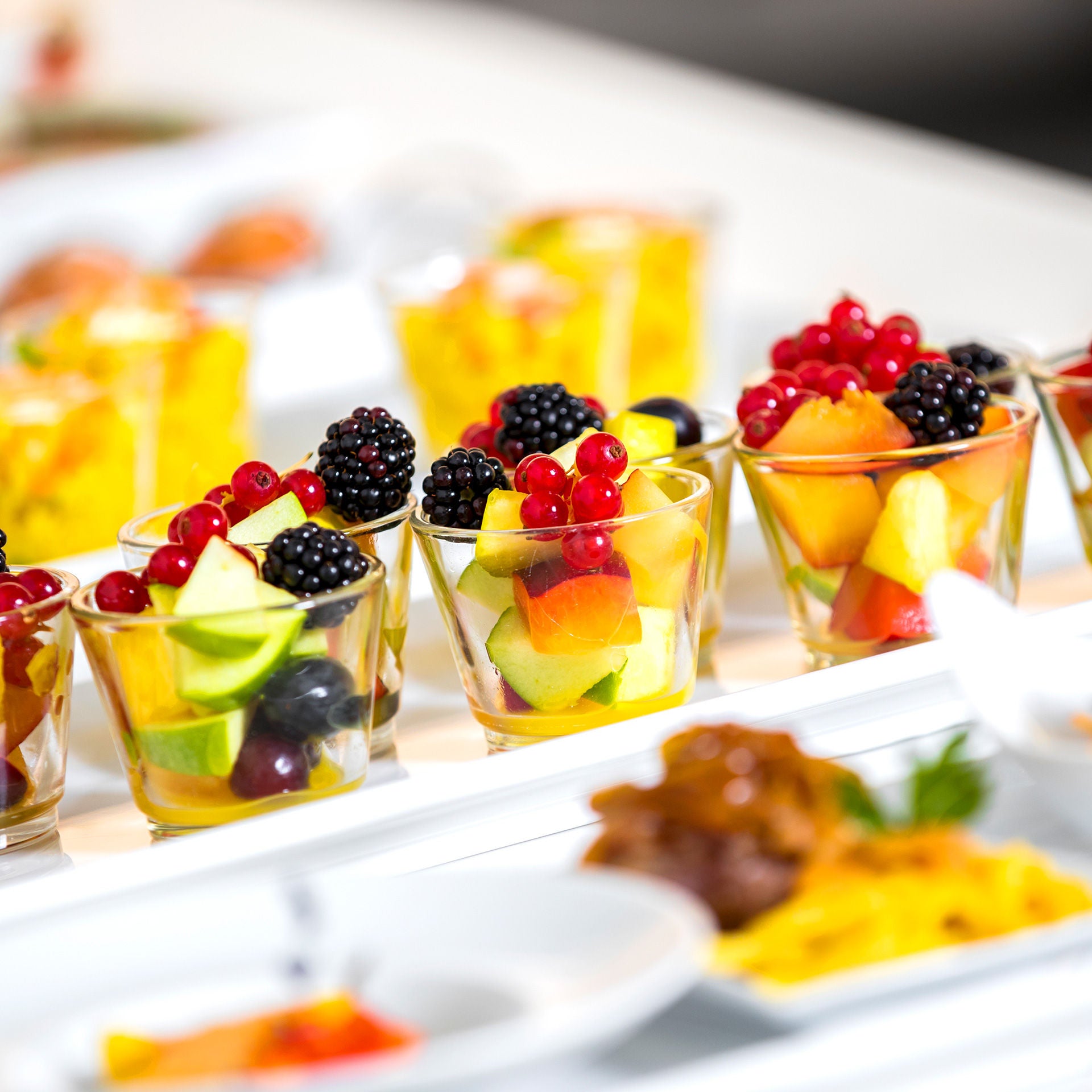 Drei Gläser mit buntem Obstsalat aus Beeren und Fruchtstücken auf einer Servierfläche.