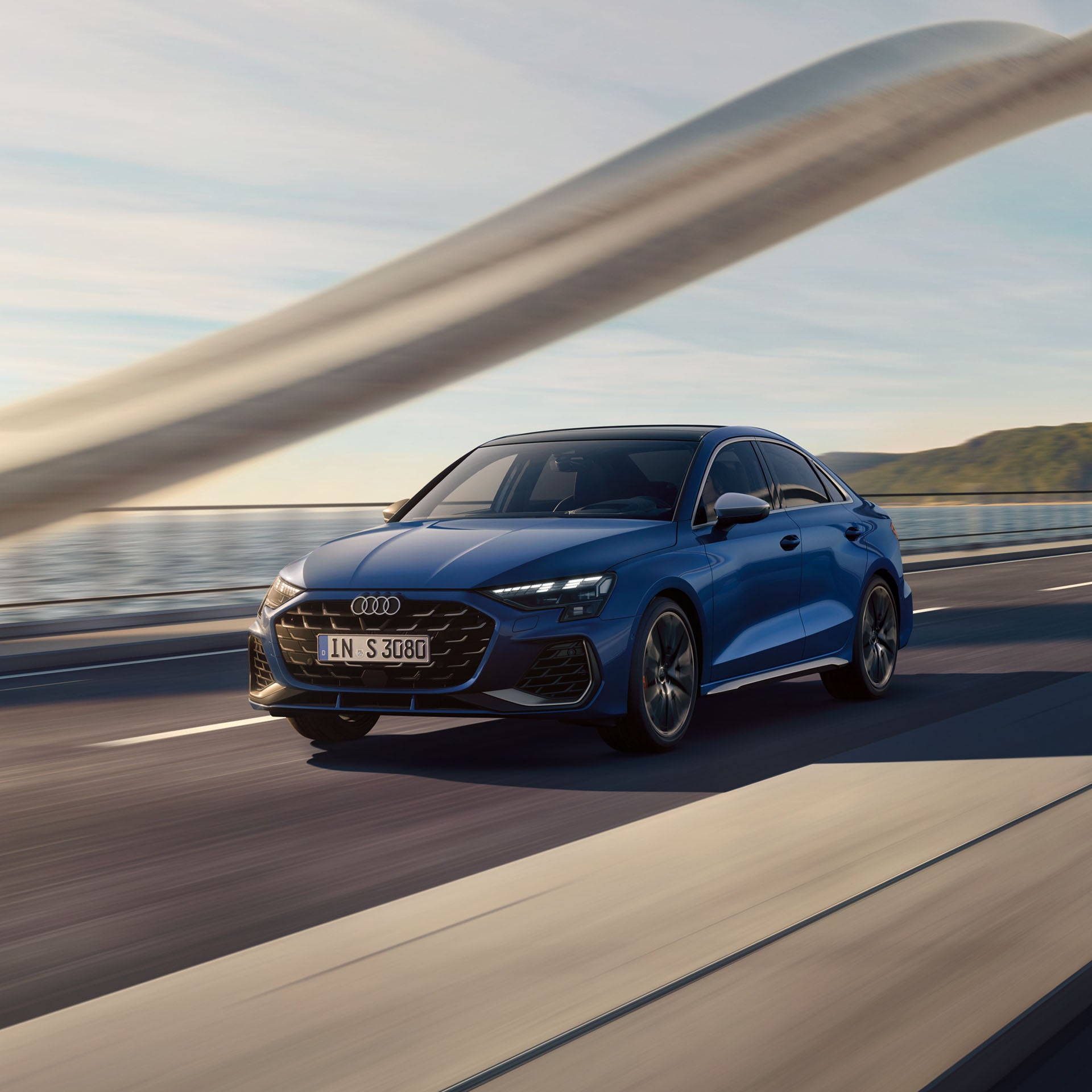 Blaue Audi S3 Limousine fährt bei Sonnenlicht auf einer Brücke am Wasser entlang, aufgenommen aus der Front-Seitenperspektive.