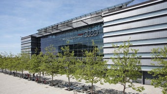 Moderne Glasfassade des Audi-Firmensitzes mit Logo und Slogan „Vorsprung durch Technik“, davor eine Allee mit Bäumen.