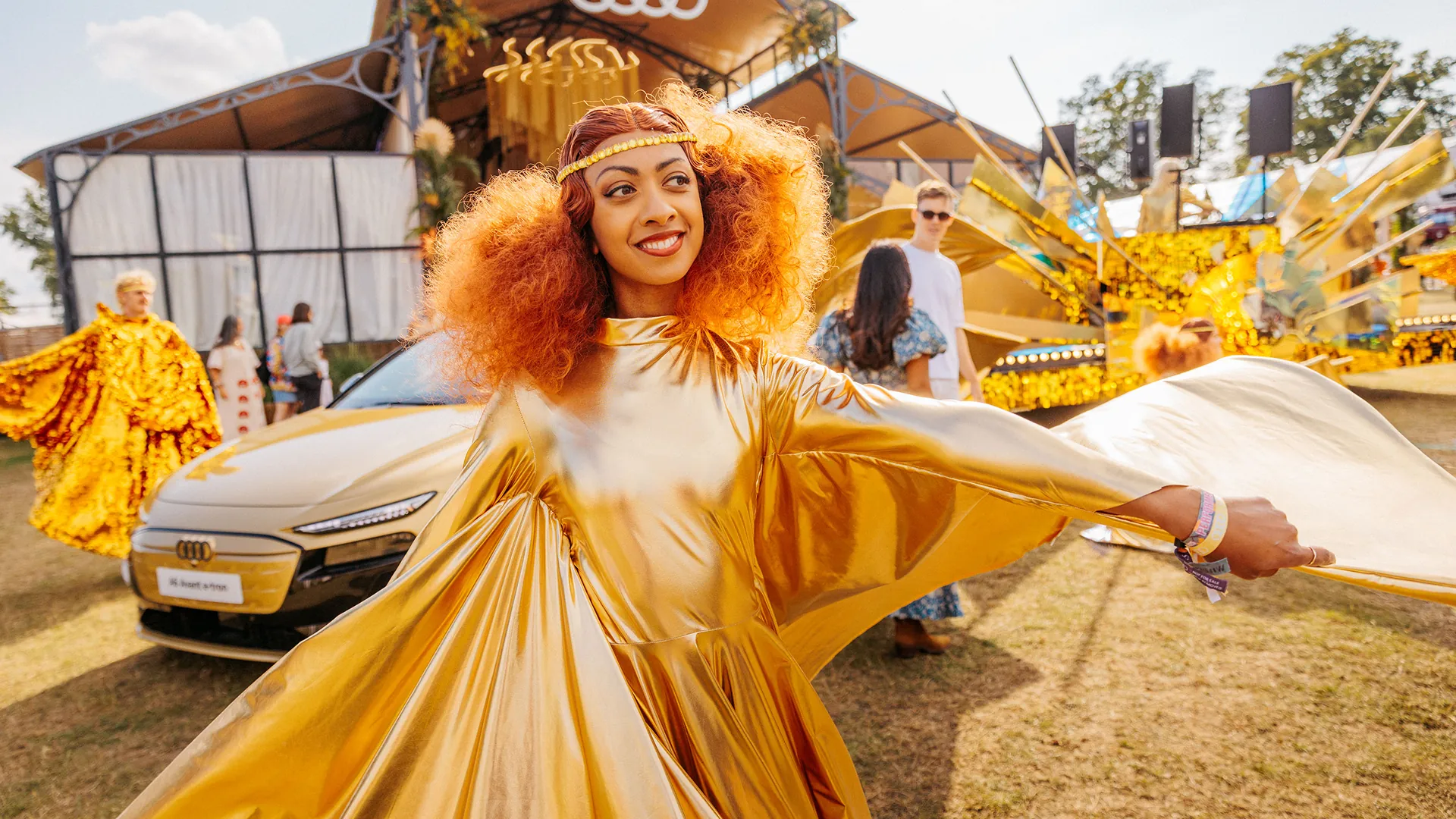 A woman with a voluminous red afro twirls in a flowing gold cape dress, with a gold Audi and the festival pavilion in the background. A woman with a voluminous red afro twirls in a flowing gold cape dress, with a gold Audi and the festival pavilion in the background.