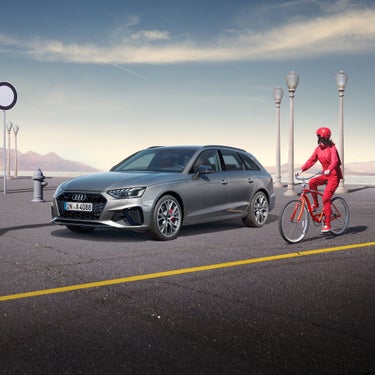 Silver car parked by a curb with a person in red on a bicycle passing by, street lamps and mountains in the background.