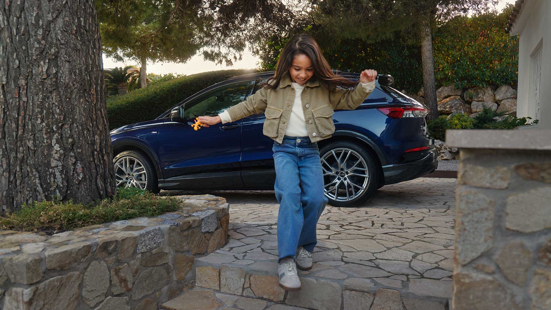Ein Kind hüpft fröhlich eine Steintreppe umgeben von Bäumen hinunter. Im Hintergrund ist ein blaues Audi-Modell zu sehen.