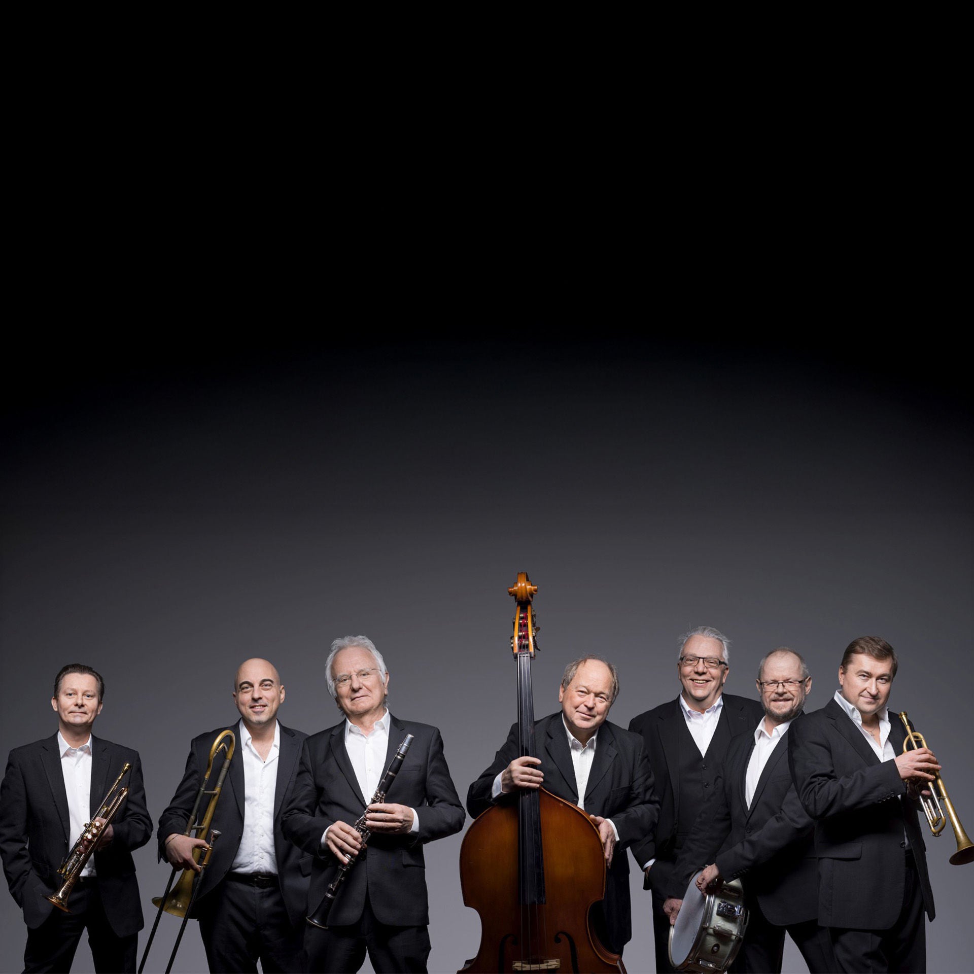 Sieben Musiker in schwarzen Anzügen stehen mit Trompete, Posaune, Klarinette, Kontrabass und Schlagzeug vor grauem Hintergrund.
