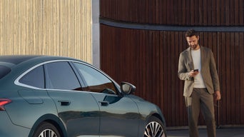 Mann mit Smartphone steht neben einem geparkten, dunkelgrünen Audi, im Hintergrund moderne Holz- und Betonarchitektur.