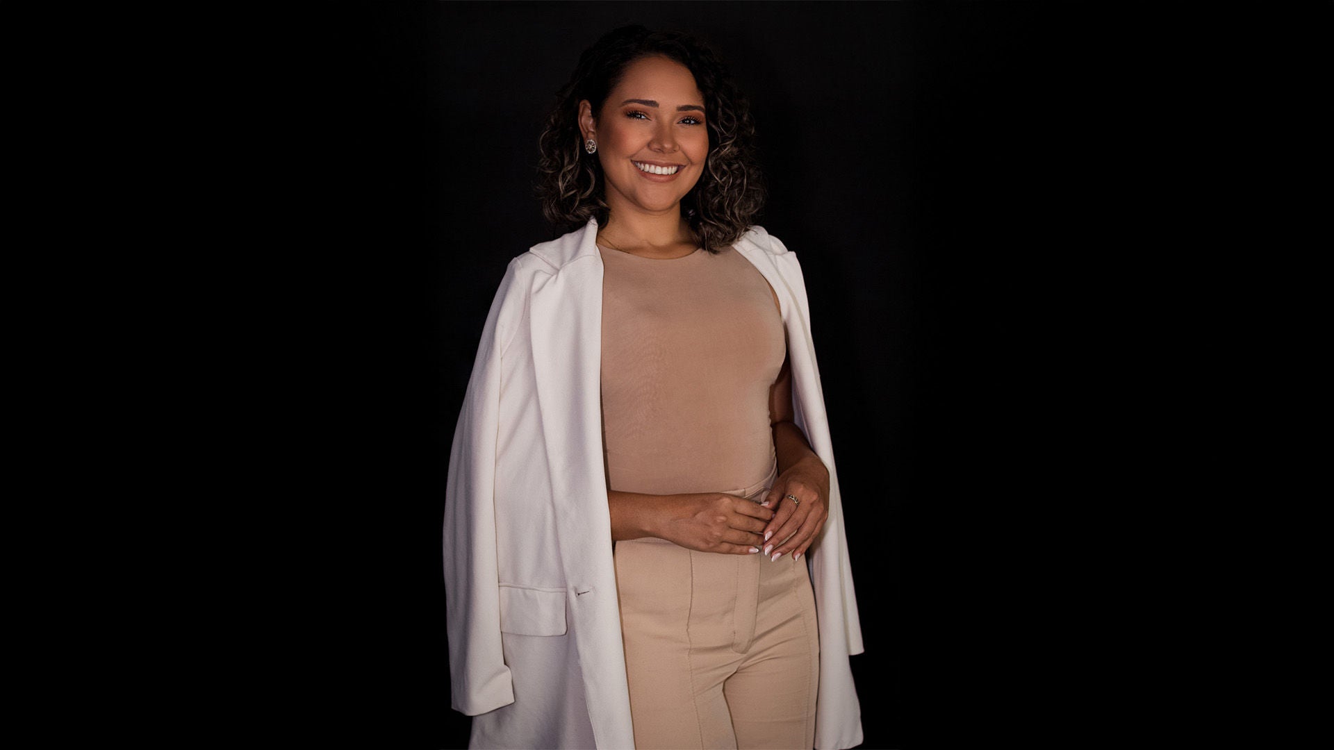 Frau im beigefarbenen Oberteil und weißem Blazer, lächelnd, vor schwarzem Hintergrund.  