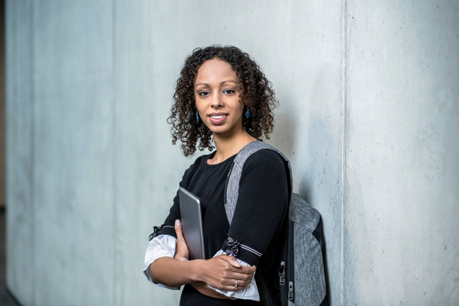 Eine lächelnde Frau mit lockigem Haar und Rucksack hält ein Tablet und lehnt an einer grauen Wand.