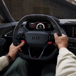 Duas mãos masculinas seguram o volante do Audi RS e-tron GT performance. O polegar esquerdo pressiona o botão vermelho de impulso.