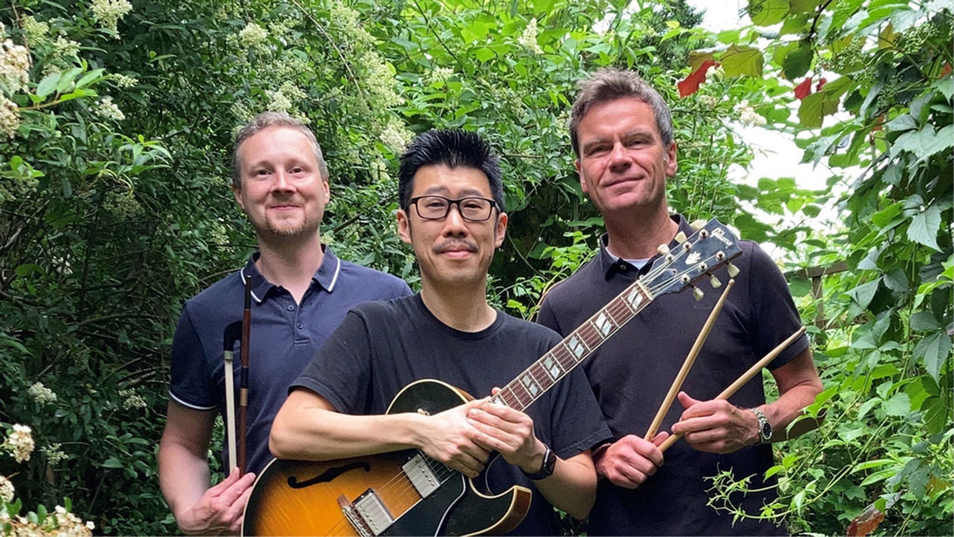 Drei Männer im Grünen; einer hält E-Gitarre, die anderen halten Drumsticks, alle schauen in die Kamera.