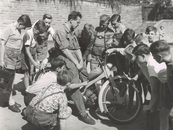 Schwarz-weiße Fotoaufnahme eines Mannes auf einer NSU Quickly, der umgeben ist von interessierten Jungen