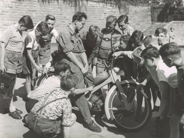 Black and white photo of a man on an NSU Quickly surrounded by interested boys