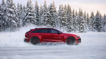 A red Audi RS 5 Avant driving on snow; snow dust, snow-covered fir trees, and a bright sky in the background.