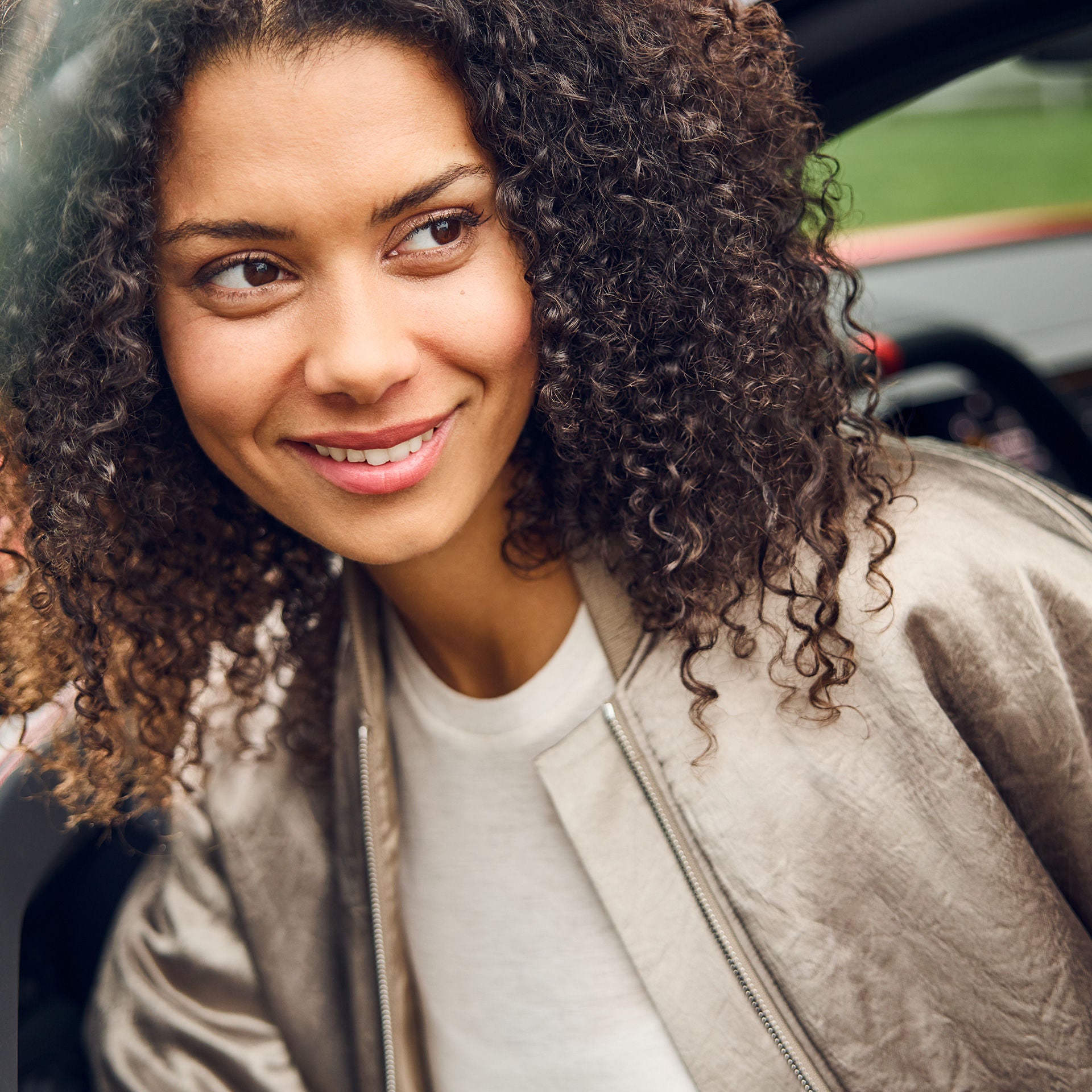 Young woman with dark, shoulder-length curls smiling, standing near an open car.