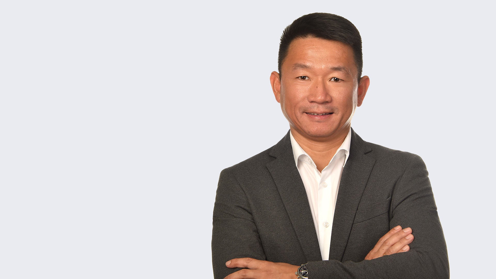 Mann im dunklen Sakko und weißem Hemd steht mit verschränkten Armen vor hellem Hintergrund und lächelt leicht.