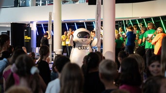 Maskottchen in weißem Audi-Kostüm steht vor Publikum; dahinter Musiker mit Blasinstrumenten in heller Innenhalle.