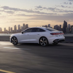 De Audi A5  e-hybrid in zijprofiel rijdend over een weg langs een waterpartij. Op de achtergrond is de skyline van een stad bij zonsopgang te zien.