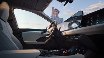 Blick in modernen Audi-Innenraum mit Touchscreen, Ambientebeleuchtung und Frau vor der Frontscheibe bei Sonnenuntergang.