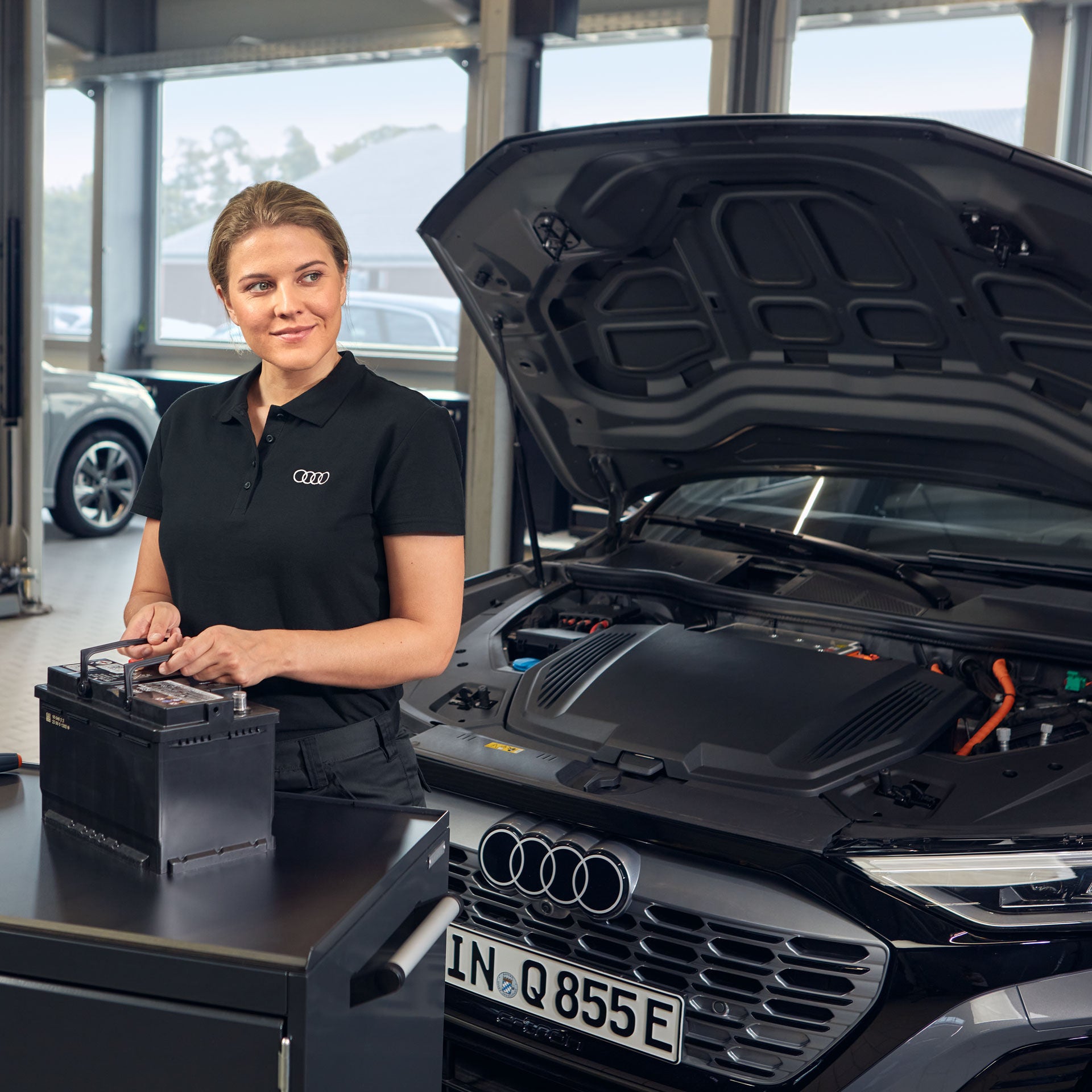 Audi-Mitarbeiterin prüft eine Autobatterie neben einem Audi mit geöffneter Motorhaube in der Werkstatt.