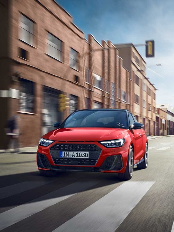 Roter Audi A1 Sportback frontal in Fahrt auf einer städtischen Straße mit Zebrastreifen und Gebäuden.