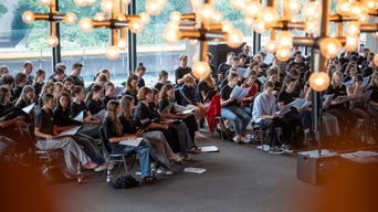 Große Gruppe junger Menschen sitzt in einem hellen Raum und probt konzentriert mit Notenblättern in der Hand.