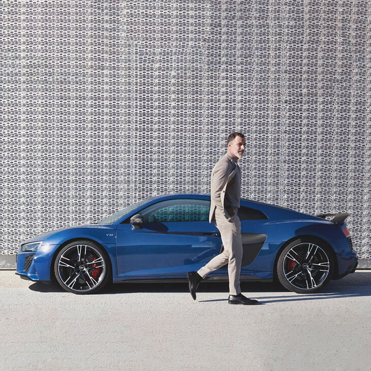 Man in a suit walking past a blue car parked in front of a textured wall.