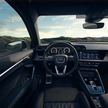 Blick vom Fahrersitz auf das Cockpit eines Audi, mit Lenkrad, digitalen Anzeigen und Infotainmentsystem vor Naturkulisse.