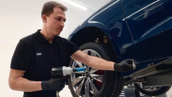 Mechaniker in schwarzer Audi-Uniform zieht Radbolzen an blauem Fahrzeug fest.