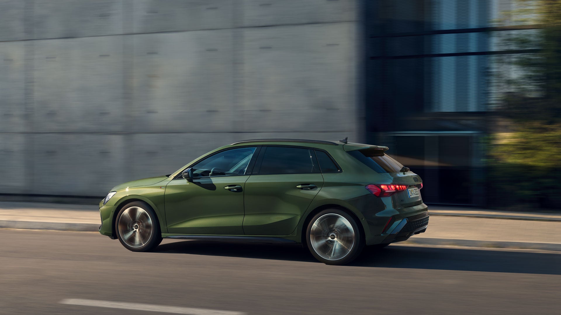 Grüner Audi A3 fährt auf einer Straße vor verschwommener moderner Gebäudefassade.