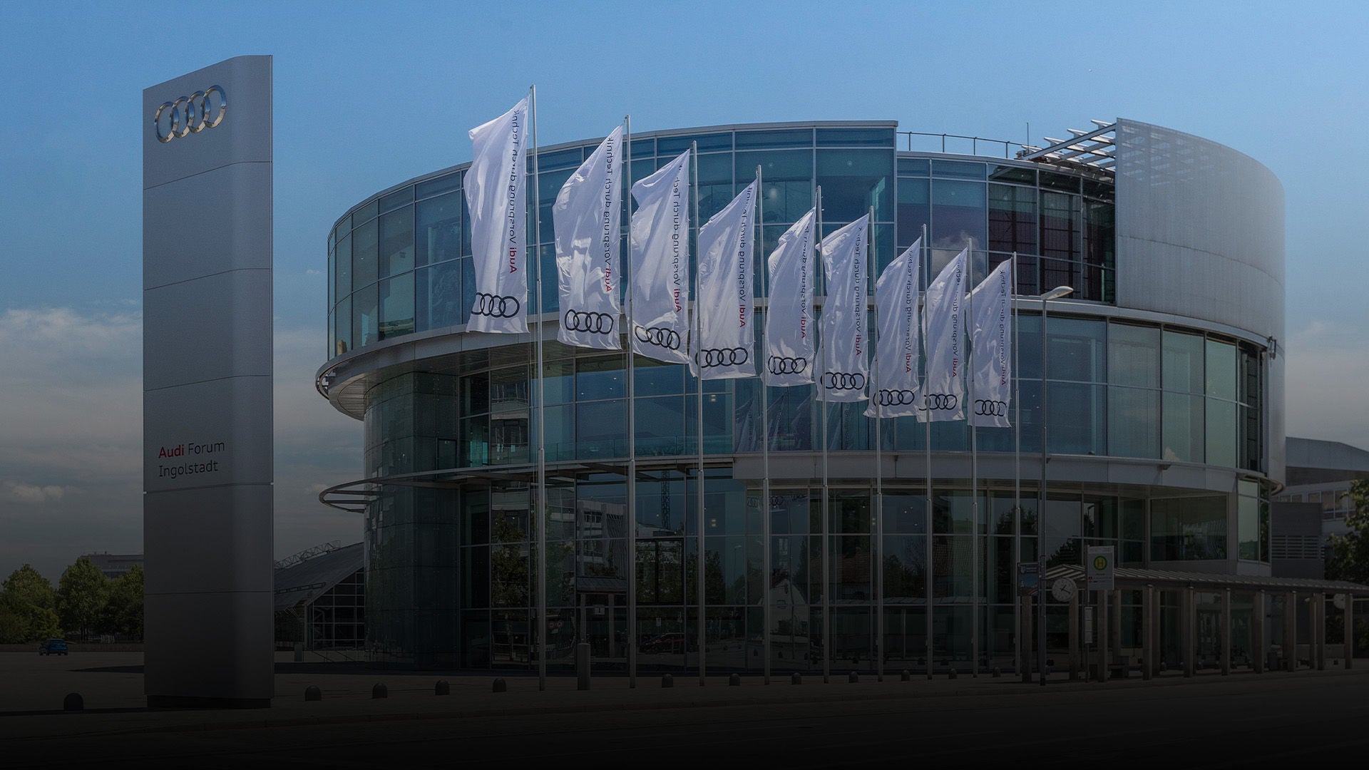 Modernes Glasgebäude des Audi Forum Ingolstadt mit Audi-Flaggen und großem Logoschild vor blauem Himmel.
