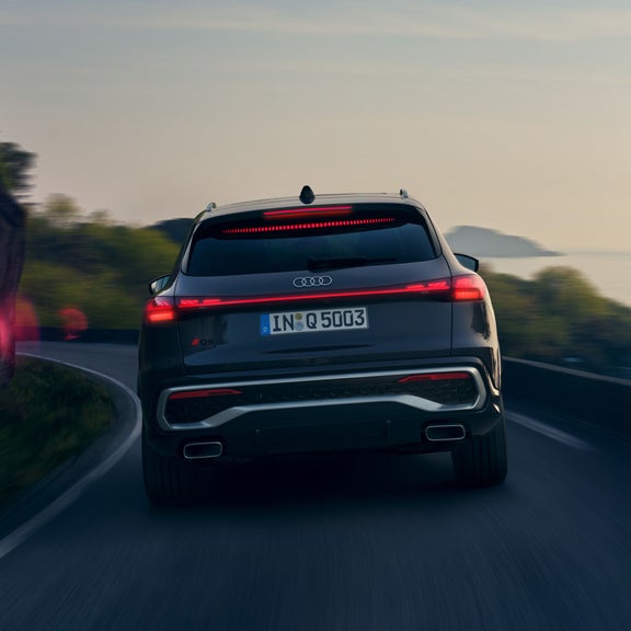 Audi SUV driving on a coastal road at dusk, with taillights on.