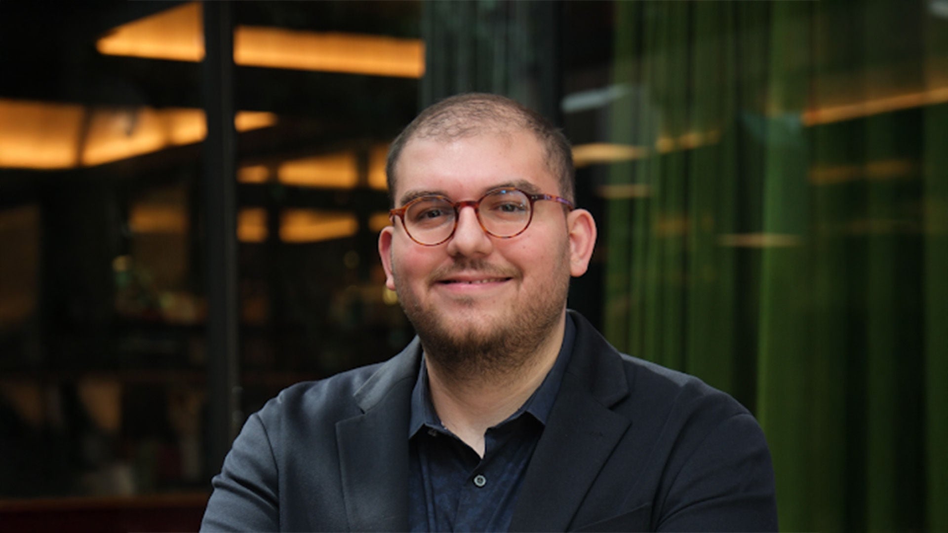 Porträt eines Mannes mit Brille im dunklen Anzug, leicht lächelnd, vor Glasfassade mit grünem Vorhang.