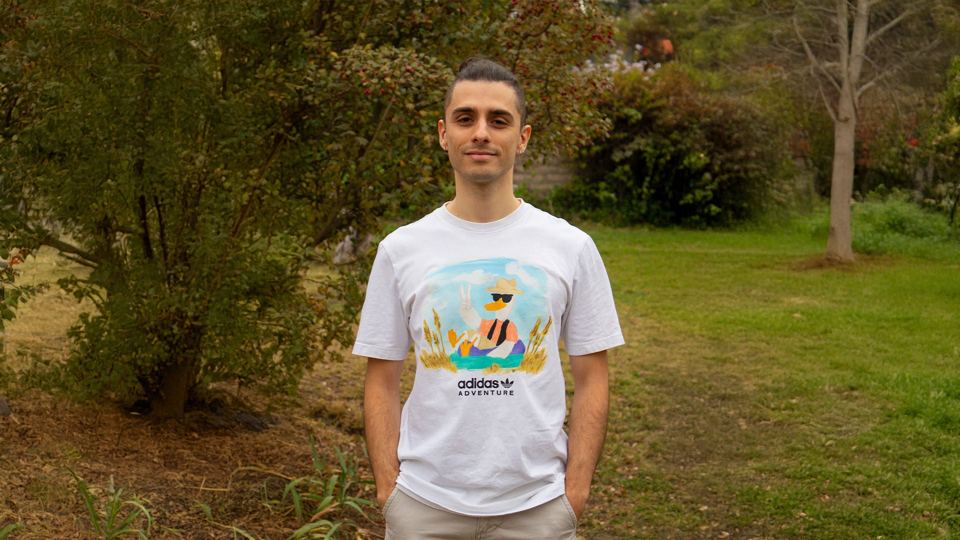 Junger Mann im weißen T-Shirt mit buntem Aufdruck, steht im Park vor Bäumen und Sträuchern.