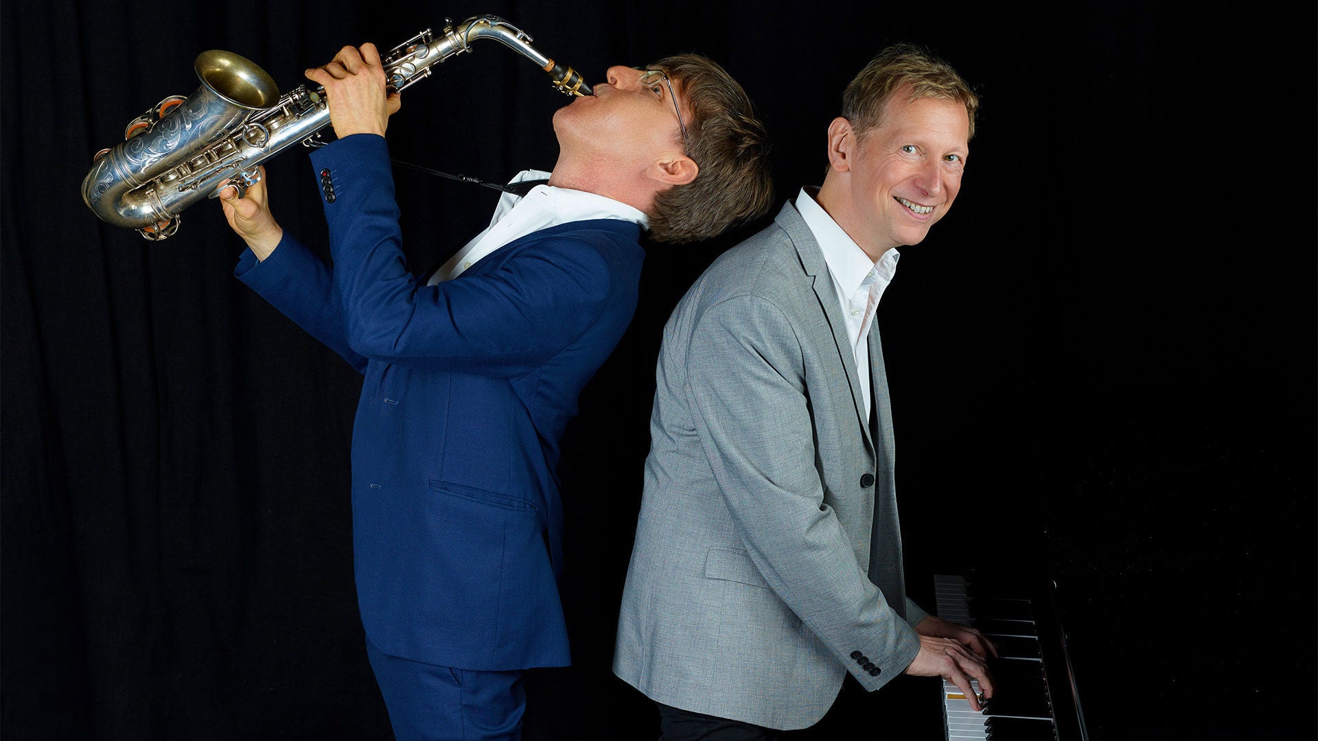 Zwei Musiker Rücken an Rücken: Saxophonist spielt nach oben, Pianist lächelt am Klavier vor schwarzem Hintergrund.