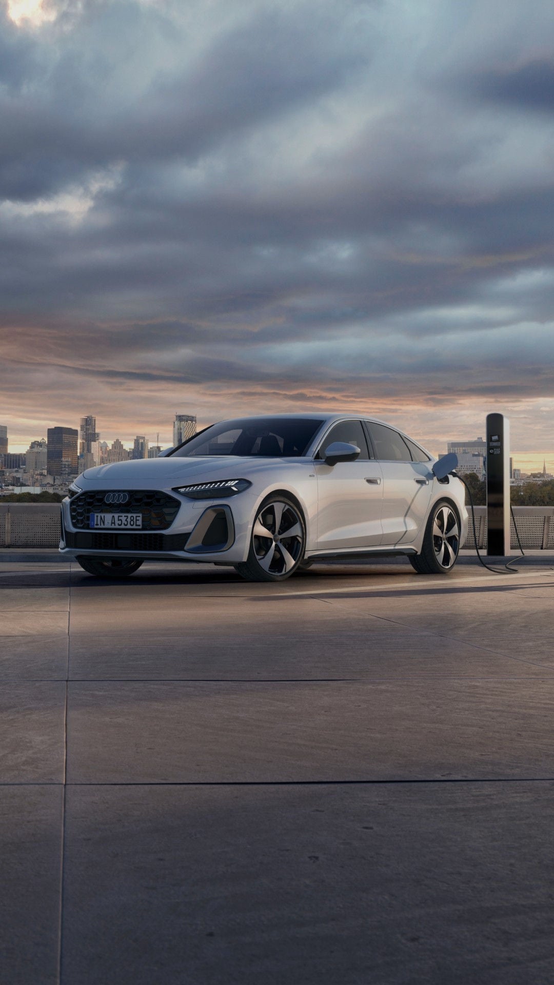 A white Audi electric car charges at a station on a rooftop parking lot. A person in a tan coat walks by, with a cityscape in the background under a cloudy sky.
