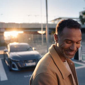  Mann im braunen Mantel steht lächelnd vor einem geparkten Audi bei Sonnenuntergang.