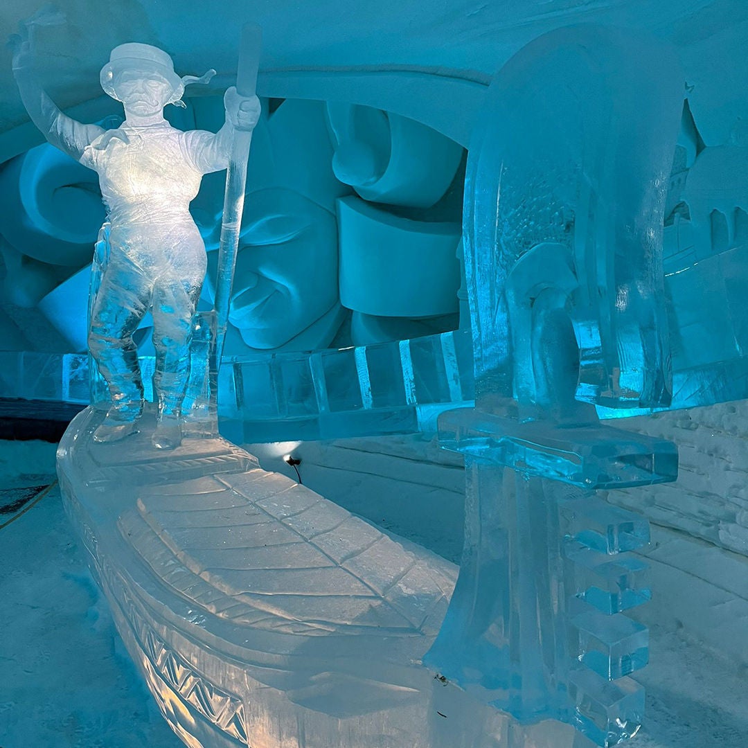 An ice sculpture of a person wearing a hat and holding a staff in a blue-lit ice chamber; to the right, a piano made of ice.