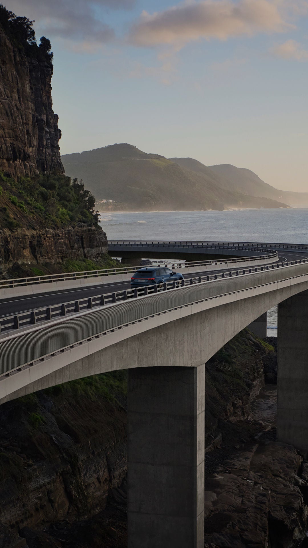 A blue car drives along a winding coastal road, flanked by cliffs and mountains under a cloudy sky. The ocean glimmers in the background.