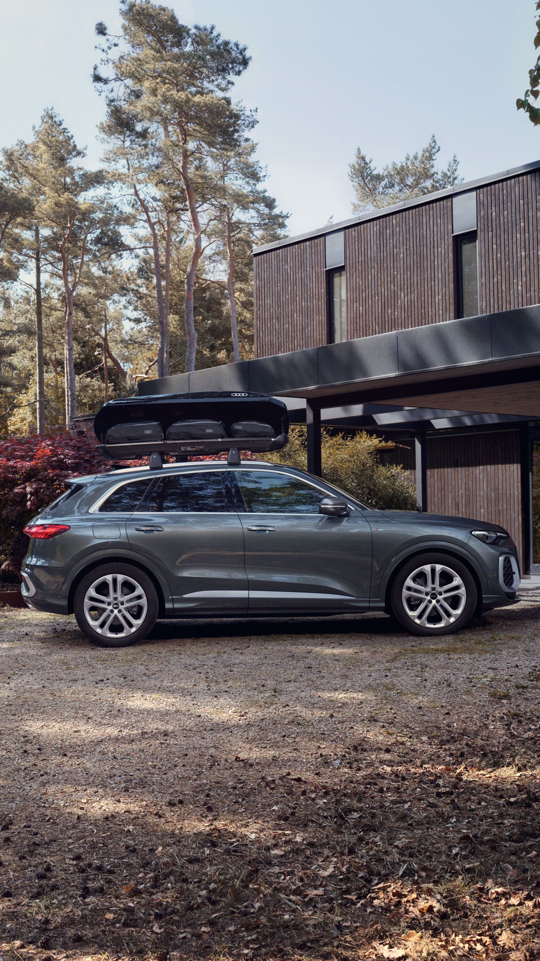 Grauer Audi SUV mit Dachbox steht vor modernem Haus im Wald.