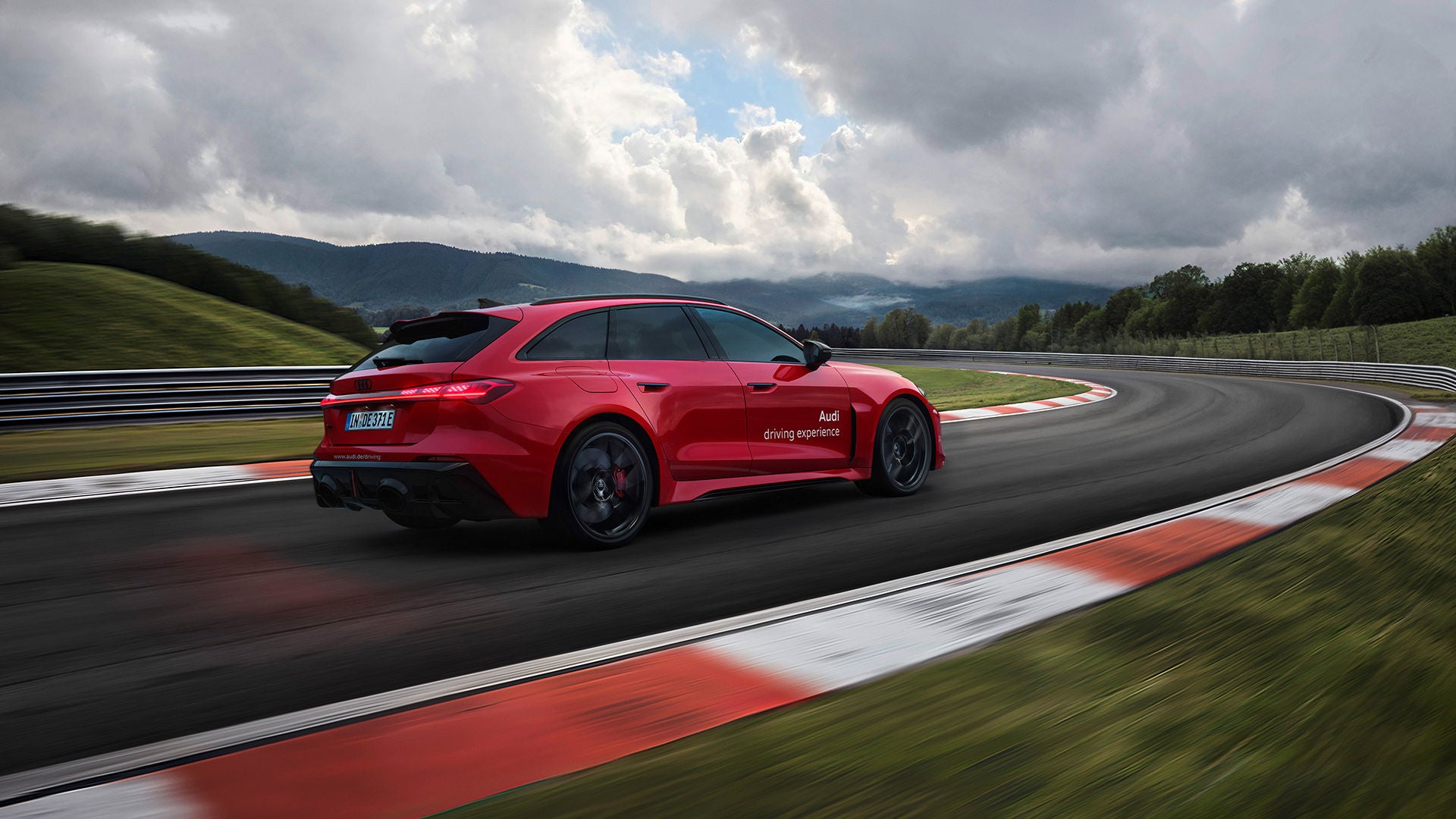 Roter Audi RS 5 Avant fährt durch eine Kurve auf der Rennstrecke; Leitplanken, grüne Hügel und wolkiger Himmel.