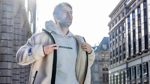 Mann in hellem Hoodie und cremefarbener Fleecejacke zeigt auf den Aufdruck „FOR THE EPIC OR EVERYDAY“.