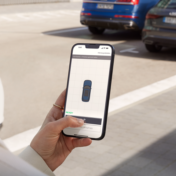 A fashionably dressed young woman is holding a smartphone, on which a stylised Audi can be seen from above. She is standing in front of parked cars.