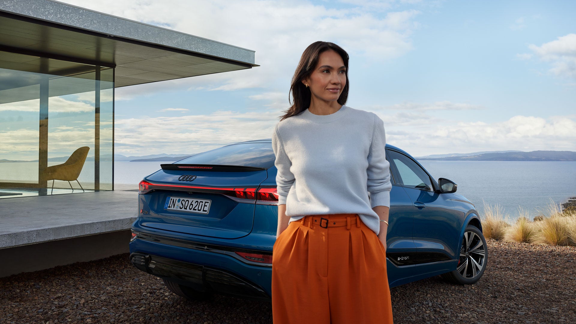 Frau steht vor einem blauen Audi SQ6 e-tron Sportback, im Hintergrund modernes Haus mit Glasfassade und Blick auf Wasser und Berge.