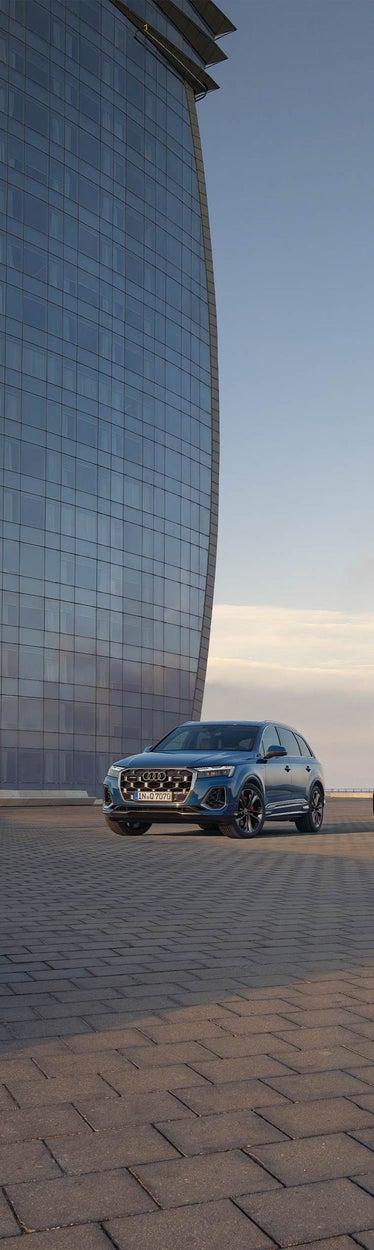 Ein blauer Audi Q7 SUV steht auf gepflastertem Platz vor gläserner Hochhausfassade, Himmel im Abendlicht.