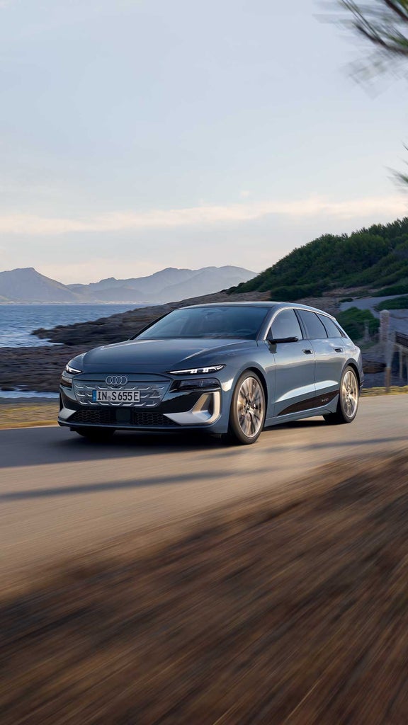 Dunkelblauer Audi S6 Avant e-tron fährt auf einer Küstenstraße mit Meer und Bergen im Hintergrund.