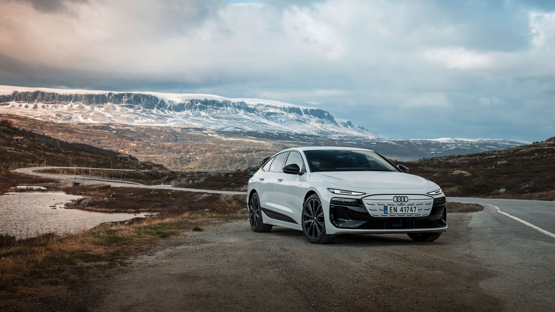 Weißer Audi A6 Sportback e-tron vor einer skandinavischen Naturkulisse mit Nadelwäldern, schneebedeckten Bergen und einem Flusslauf.