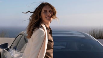 Smiling woman with wavy hair stands beside a silver car, wearing a white blazer and brown top. Ocean view in background, conveying a relaxed, joyful vibe.