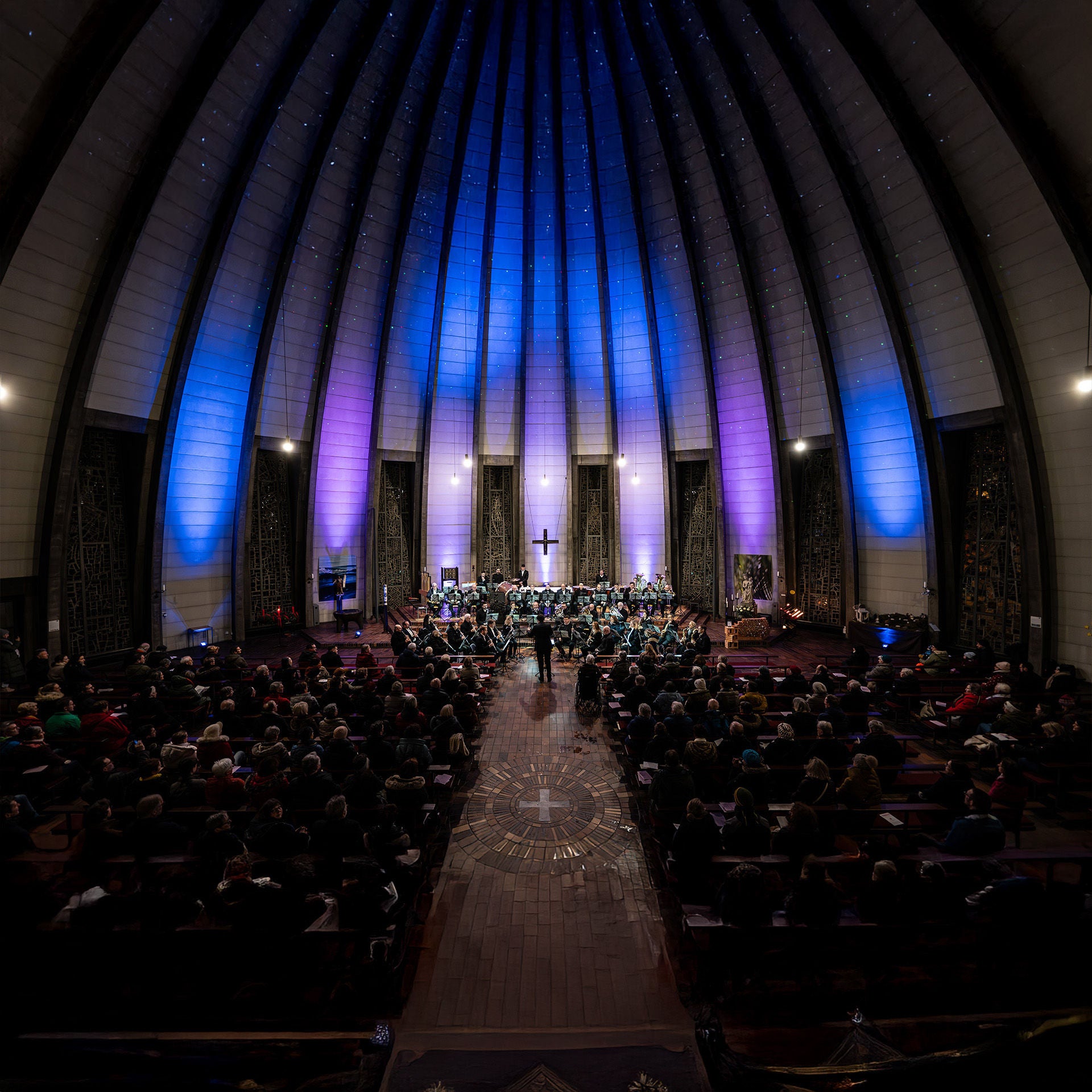 Blick in eine Kirche mit hoher, beleuchteter Decke, ein Orchester spielt vor einem sitzenden Publikum.