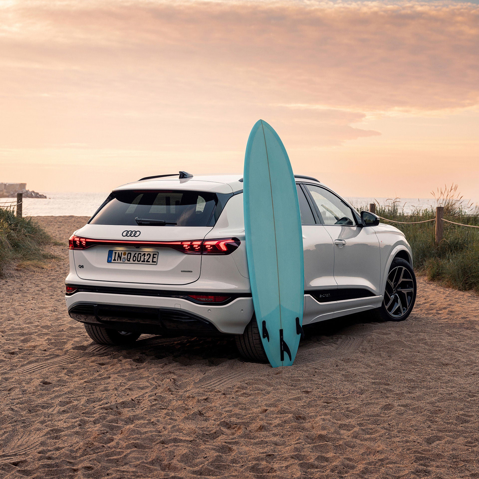 Ein weißer Audi Q6 SUV e-tron parkt am Strandübergang, im Hintergrund das Meer. Ein türkises Surfbrett lehnt an der hinteren Seitentür.