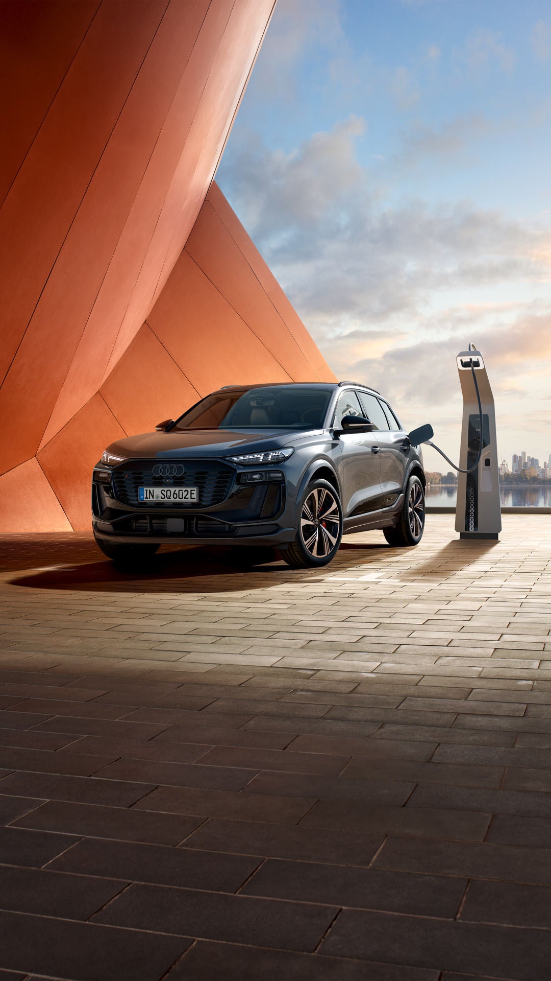 Seitliche Front des SQ6 SUV e-tron, welcher neben einer orangenen Gebäudefassade an eine Ladestation angeschlossen ist. Im Hintergrund ist das Meer, sowie die Skyline einer Stadt zu erkennen.